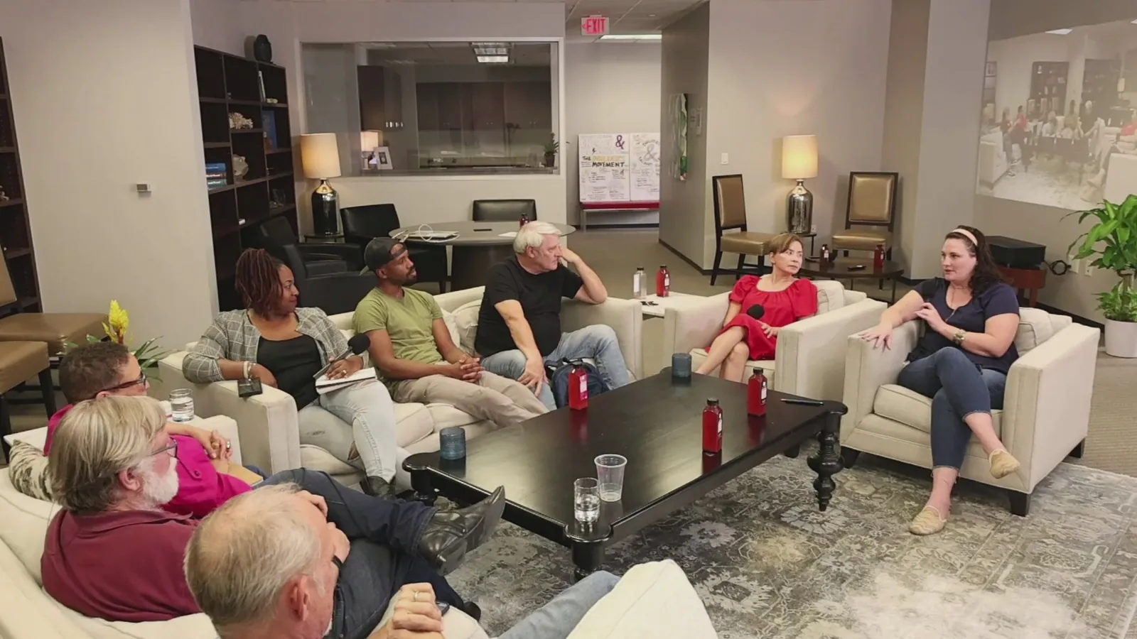 A woman in an armchair at the end of a coffee table, with a circle of people seated on couches around her, listening